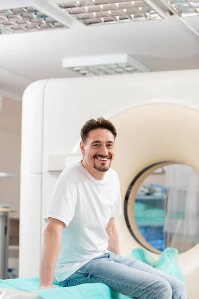 Cheerful brunette man in white t-shirt sitting near ct scanner and smiling at camera — Stock Photo
