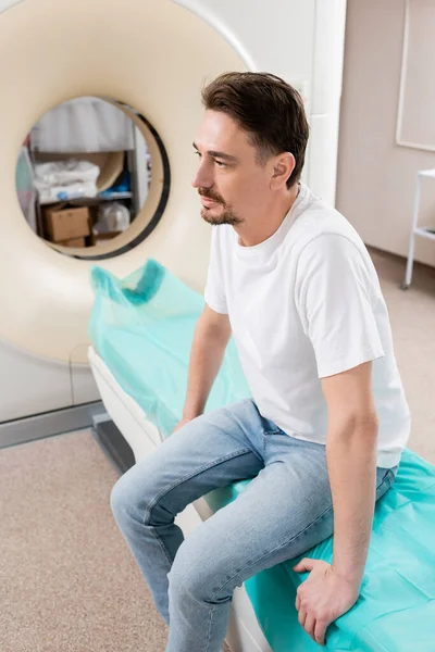 Nachdenklicher Mann schaut weg, während er neben Computertomographen im Krankenhaus sitzt — Stockfoto