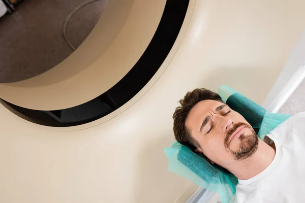 High angle view of tense brunette man with closed eyes doing diagnostics on computed tomography scanner — Stock Photo