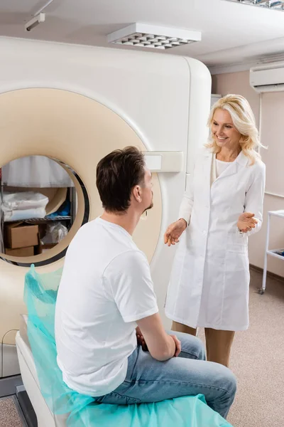 Radiologista loira sorridente falando com homem morena sentado perto do scanner de tomografia computadorizada — Fotografia de Stock