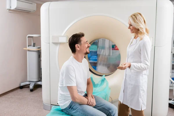 Vista lateral del radiólogo rubio hablando con el paciente sonriente cerca de la máquina de tomografía computarizada - foto de stock
