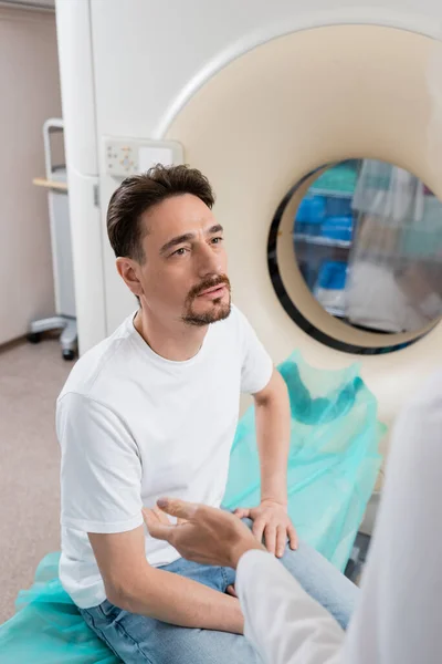 Homme assis près de la machine de tomodensitométrie et regardant le médecin au premier plan flou — Photo de stock