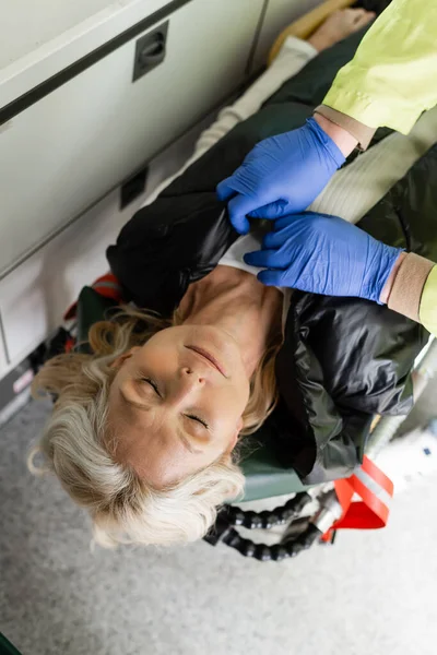 Вид сверху на парамедика снимающего куртку с бессознательной женщины средних лет в машине скорой помощи — стоковое фото