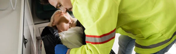 Paramédico de uniforme tirando jaqueta de mulher inconsciente borrada em veículo de emergência, banner — Fotografia de Stock