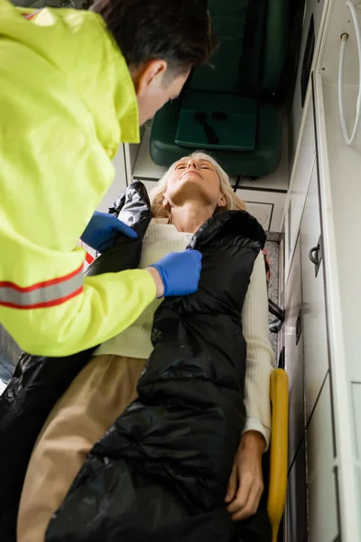 Paramédico borroso quitando la chaqueta de la mujer inconsciente de mediana edad en el vehículo de emergencia - foto de stock