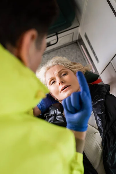 Високий кут зору розмитого парамедика, що показує палець, надаючи першу допомогу зрілій жінці в аварійному транспортному засобі — стокове фото
