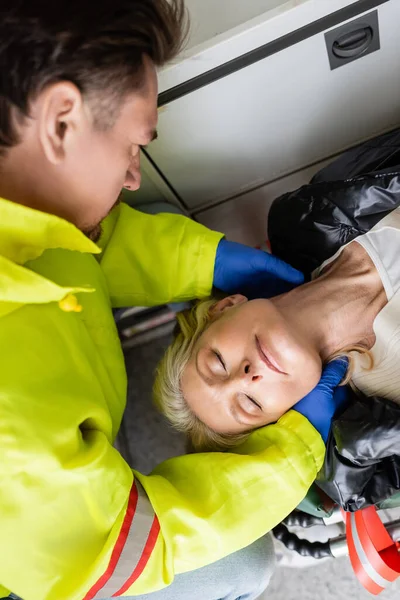 Vista superior de paramédico en guantes de látex comprobando cuello de mujer de mediana edad en vehículo de emergencia - foto de stock