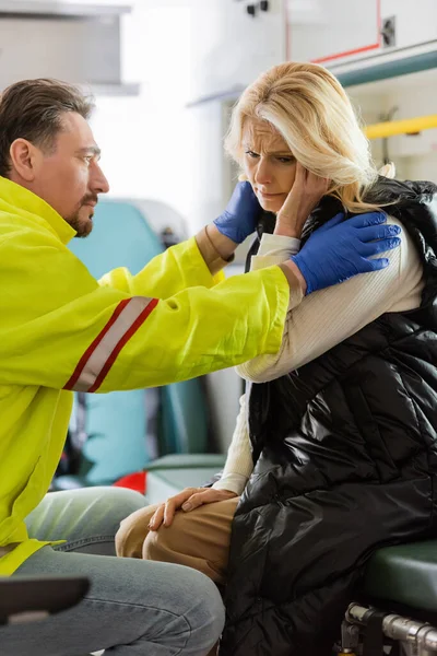 Mujer de mediana edad enferma tocando la cabeza cerca de paramédico en uniforme en vehículo de emergencia - foto de stock