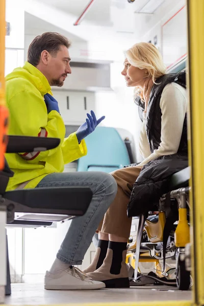 Vista lateral del gesto paramédico y hablar con la mujer de mediana edad preocupada en el vehículo de emergencia - foto de stock
