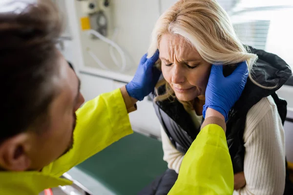 Paramédico borroso en guantes de látex revisando la cabeza de la mujer de mediana edad en el coche de emergencia - foto de stock
