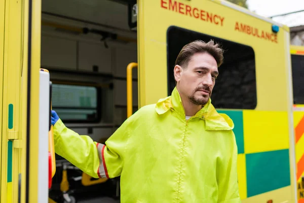 Paramédico en guante de látex y chaqueta mirando a la cámara cerca de vehículo ambulancia borrosa - foto de stock