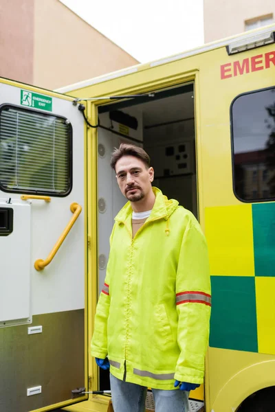 Paramedic in jacket looking at camera near ambulance car outdoors — Stock Photo