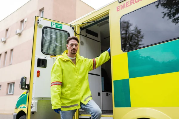 Paramédico en guantes de látex y chaqueta mirando a la cámara cerca de coche ambulancia al aire libre - foto de stock