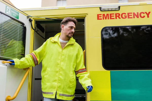 Paramédico en guantes de látex y chaqueta que abre la puerta del vehículo ambulancia al aire libre - foto de stock