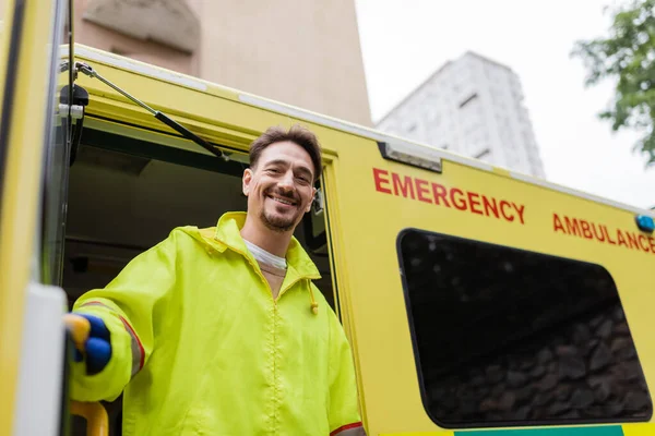 Sonriente paramédico mirando a la cámara cerca de vehículo ambulancia con puerta abierta en la calle - foto de stock