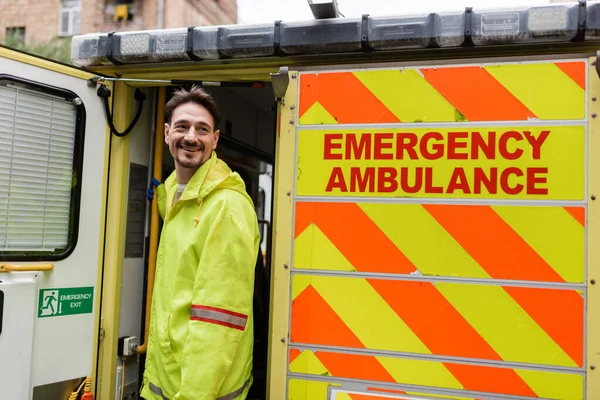 Positive paramedic looking away near ambulance car with ambulance lettering outdoors — Stock Photo