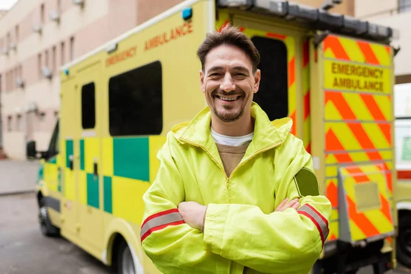 Lächelnder Sanitäter verschränkt die Arme und blickt in die Kamera in der Nähe eines verschwommenen Rettungswagens — Stockfoto