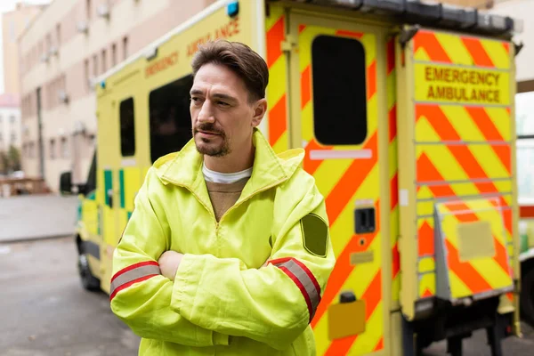 Paramedic in uniform crossing arms near blurred ambulance car outdoors — Stock Photo