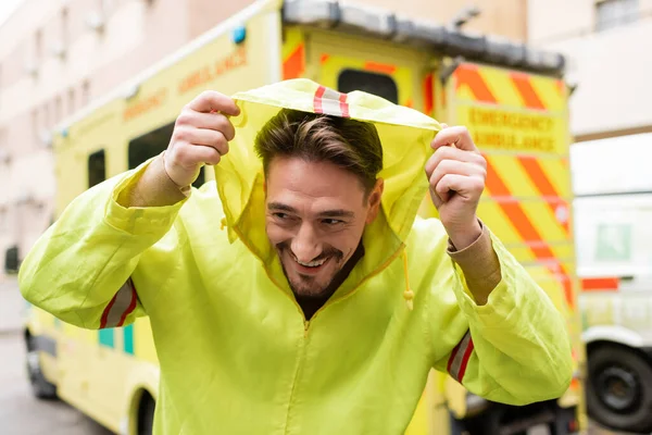 Lächelnder Sanitäter mit Kapuze in Jacke neben verschwommenem Rettungswagen im Freien — Stockfoto