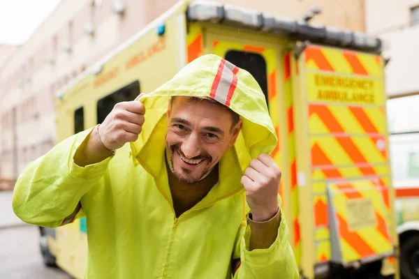 Cheerful paramedic in jacket with hood looking at camera near blurred emergency car outdoors — Stock Photo