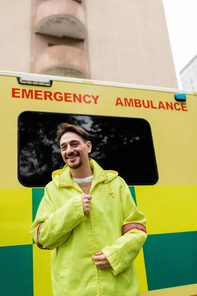 Улыбающийся парамедик в куртке и смотрящий на камеру возле машины скорой помощи на улице — стоковое фото