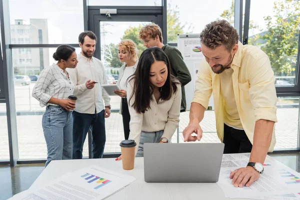 Positiver Geschäftsmann zeigt auf Laptop neben asiatischem Kollegen, während gemischtes Team über Hintergrund spricht — Stockfoto
