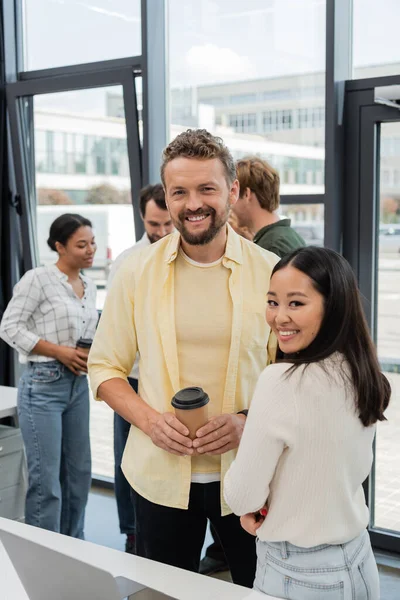 Feliz hombre de negocios con papel taza mirando cámara cerca asiático colega y equipo en fondo - foto de stock