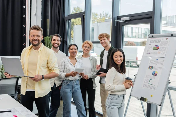 Cheerful multiethnic corporate team looking at camera near flip chart with infographics — Stock Photo