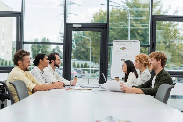 Equipe corporativa multiétnica sentada na mesa de conferência perto de flip chart com infográficos — Fotografia de Stock
