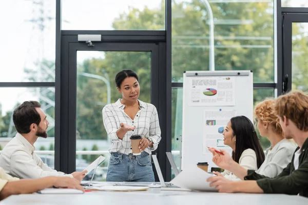 Multiracial woman with paper cup talking to colleagues during meeting — Stock Photo