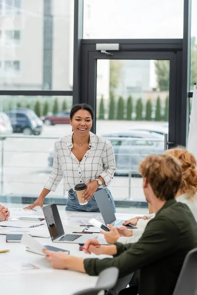 Счастливая многорасовая женщина с бумажной чашкой беседует с бизнес-командой во время встречи — стоковое фото