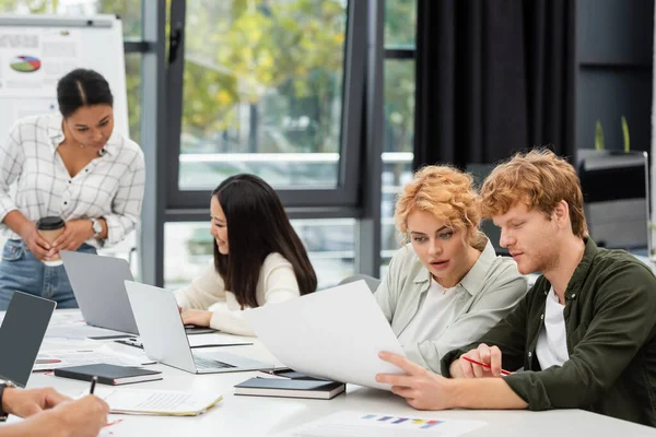 Young managers looking at document near multiethnic coworkers on blurred background — Stock Photo