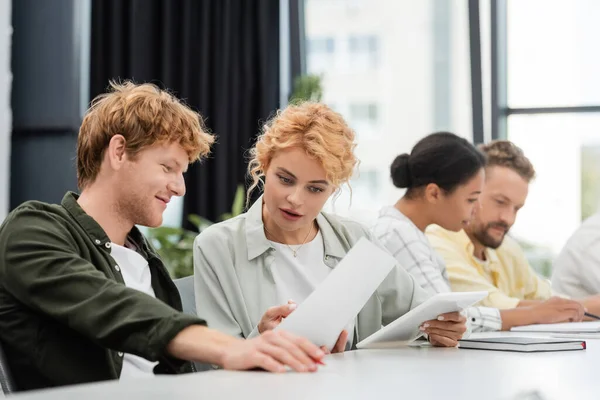 Smiling businesswoman showing document to colleague near interracial team on blurred background — Stock Photo