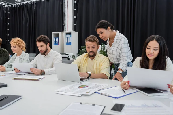 Мультирасовая женщина смотрит на ноутбук рядом с коллегой и командой, работающей за конференц-столом — стоковое фото