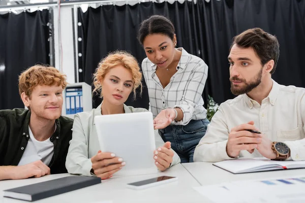 Многорасовая предпринимательница, указывающая на цифровой планшет рядом с менеджерами, работающими в офисе — стоковое фото