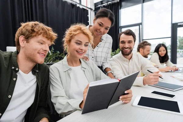 Happy businesswoman showing notebook to smiling multiethnic colleagues during meeting in office — Stock Photo