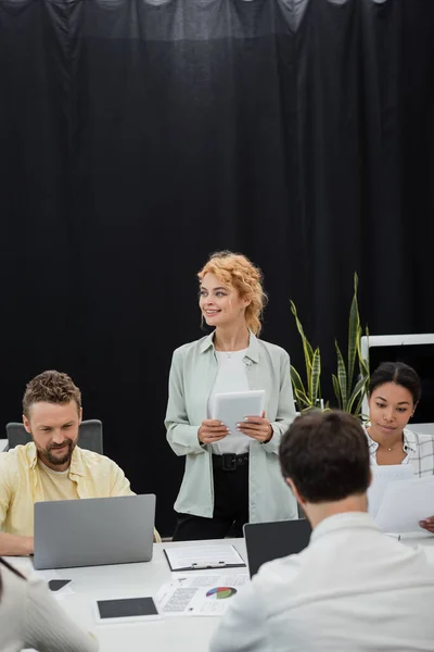Mujer de negocios rubia con tableta digital mirando hacia otro lado cerca de colegas interracial trabajando en la oficina - foto de stock