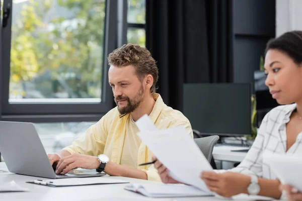 Bearded businessman typing on laptop near multiracial woman on blurred foreground — Stock Photo