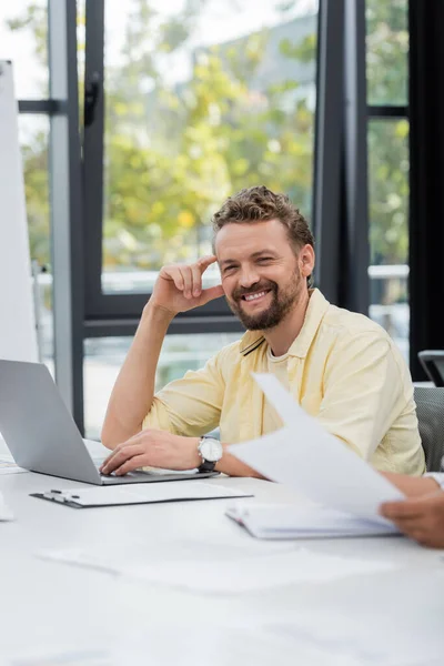 Hombre de negocios barbudo positivo sonriendo a la cámara cerca de la computadora portátil en el lugar de trabajo - foto de stock