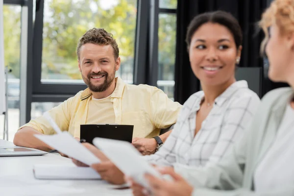 Happy bearded businessman looking at camera near interracial women talking on blurred foreground — Stock Photo