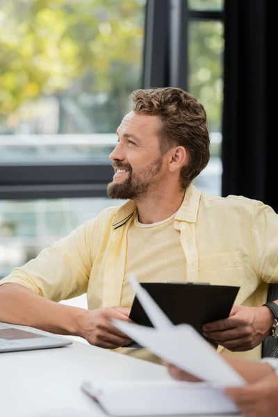 Happy bearded businessman sitting with clipboard and looking away in office — Stock Photo