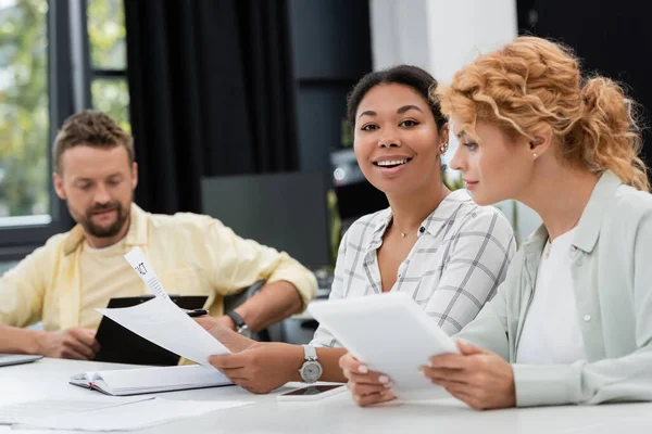 Cheerful multiracial businesswoman with papers smiling at camera near colleagues in office — Stock Photo