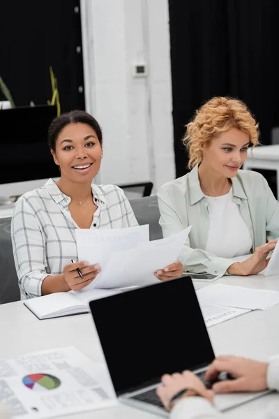 Брюнетка багаторасовий бізнес-леді тримає документи біля блондинки колеги і ноутбук з порожнім екраном на розмитому фоні — Stock Photo