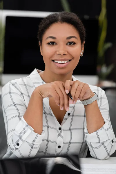 Glückliche multiethnische Geschäftsfrau hält Stift in der Hand und lächelt in die Kamera im Büro — Stockfoto