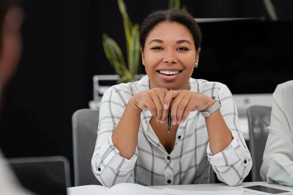 Positive multiethnische Geschäftsfrau hält Stift in der Hand und lächelt in die Kamera am Arbeitsplatz im Büro — Stockfoto