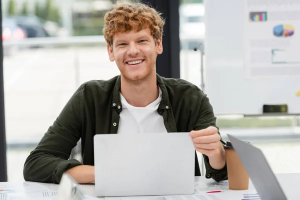 Cheerful redhead manager smiling at camera near laptop and paper cup at workplace — Stock Photo