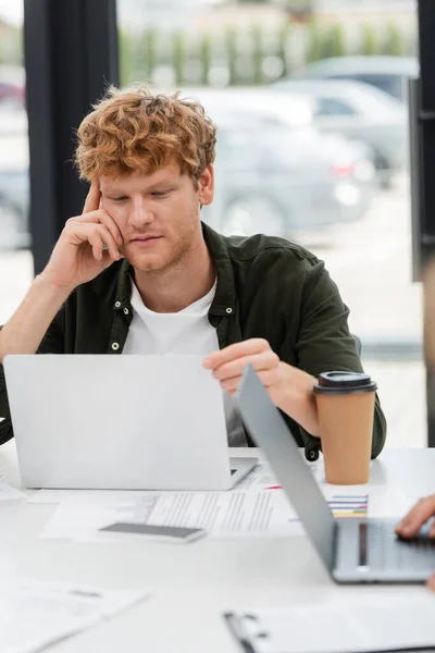 Молодой бизнесмен с рыжими волосами думает около ноутбука и кофе, чтобы пойти в офис — стоковое фото
