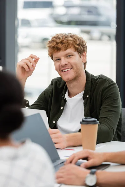 Positive redhead businessman smiling at camera near colleagues on blurred foreground — Stock Photo