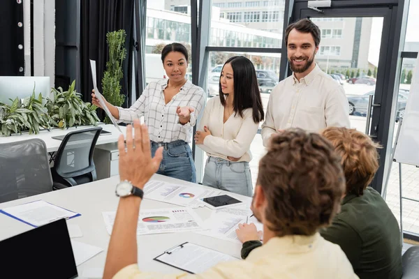 Multiracial woman holding document and pointing at colleague with raised hand on blurred foreground — Stock Photo