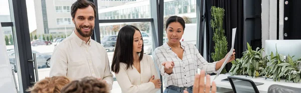 Heureuse femme multiraciale tenant document et pointant avec la main près de collègues d'affaires multiethniques, bannière — Photo de stock